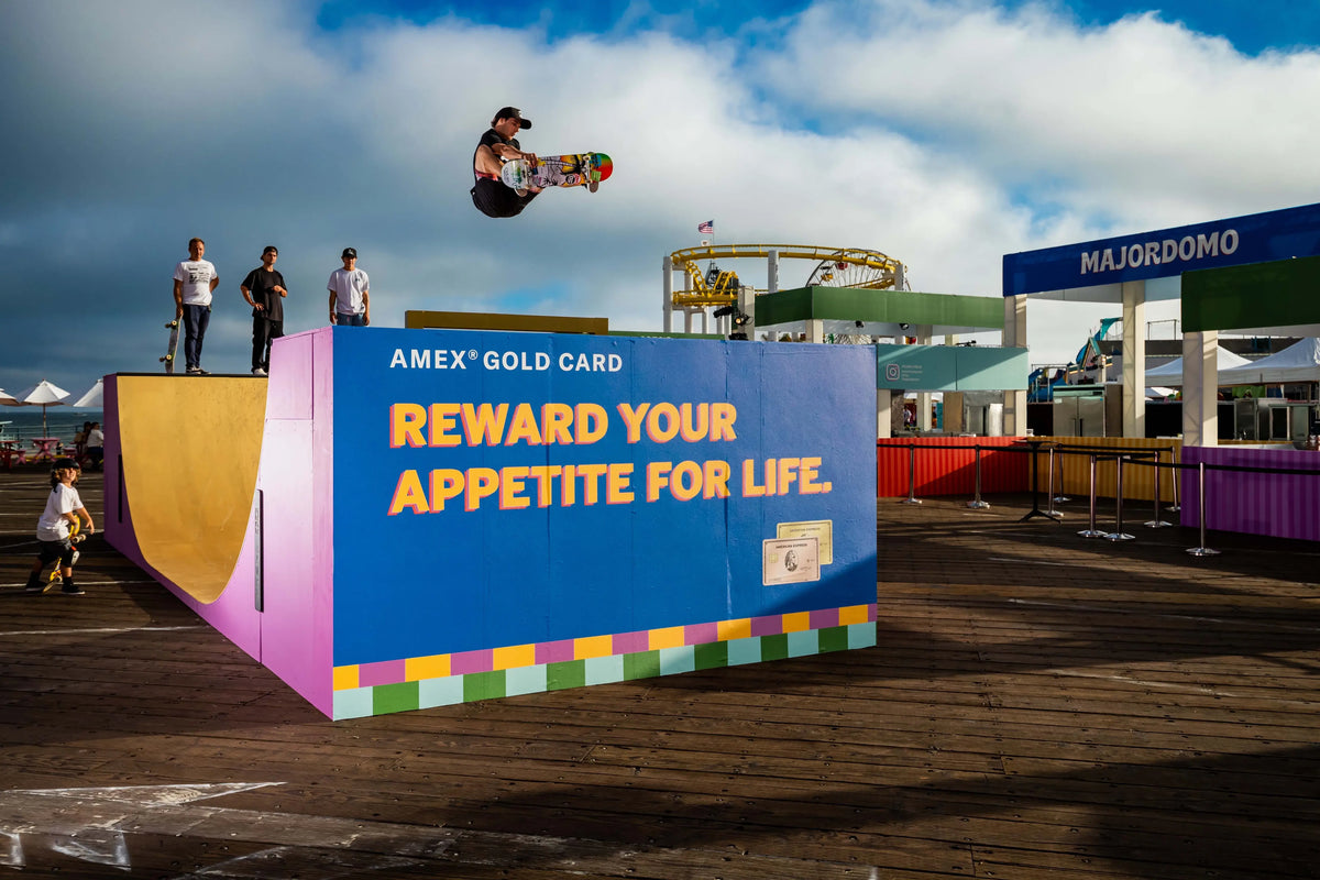 A skateboarder mid-air performs a trick in front of a large colorful ramp with the text 'AMEX GOLD CARD REWARD YOUR APPETITE FOR LIFE.' on a wooden boardwalk under a cloudy blue sky.