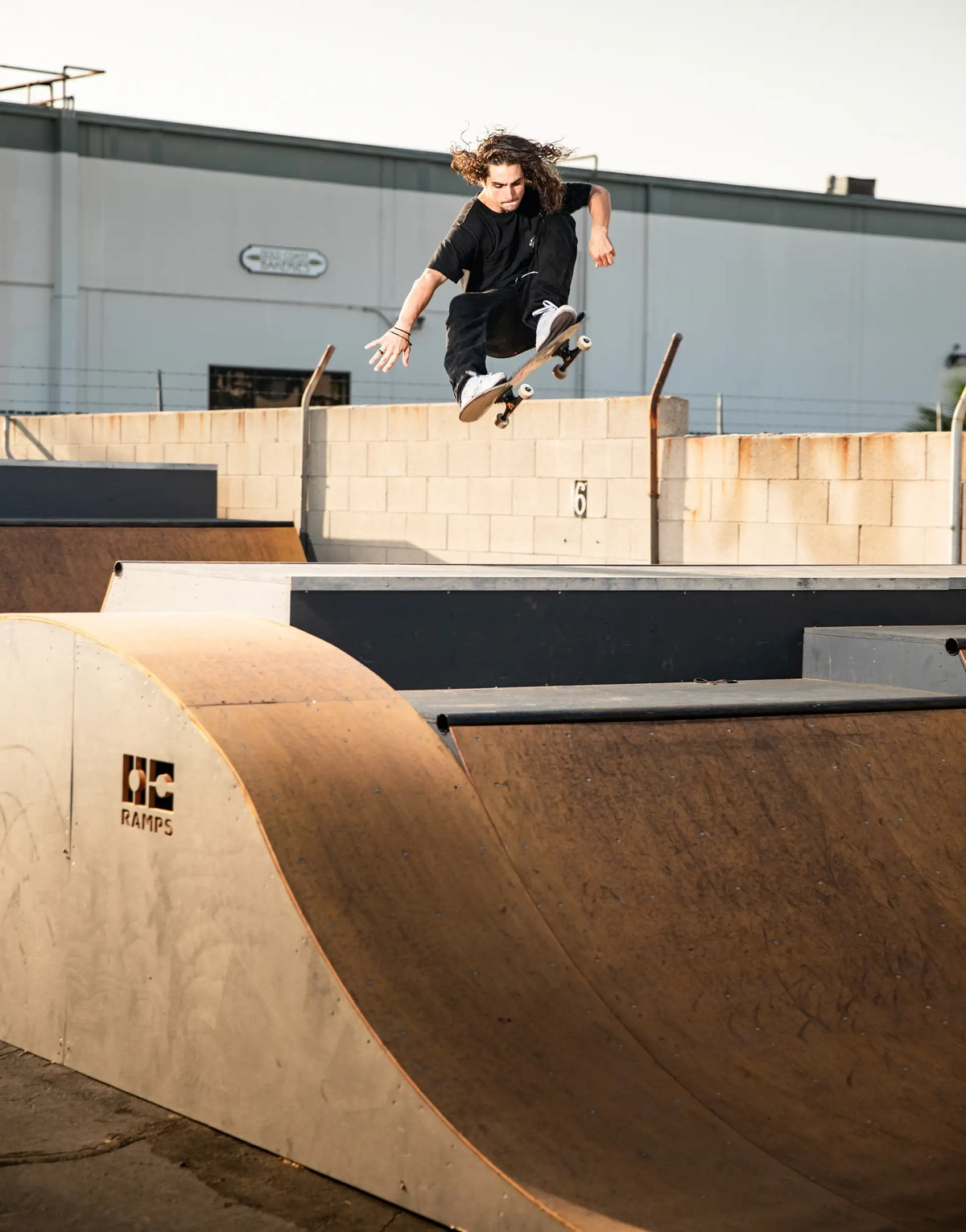 A skateboarder mid-air performing a trick at an outdoor skatepark, with a OC Ramps wooden quarter pipe in the foreground and a building in the background.