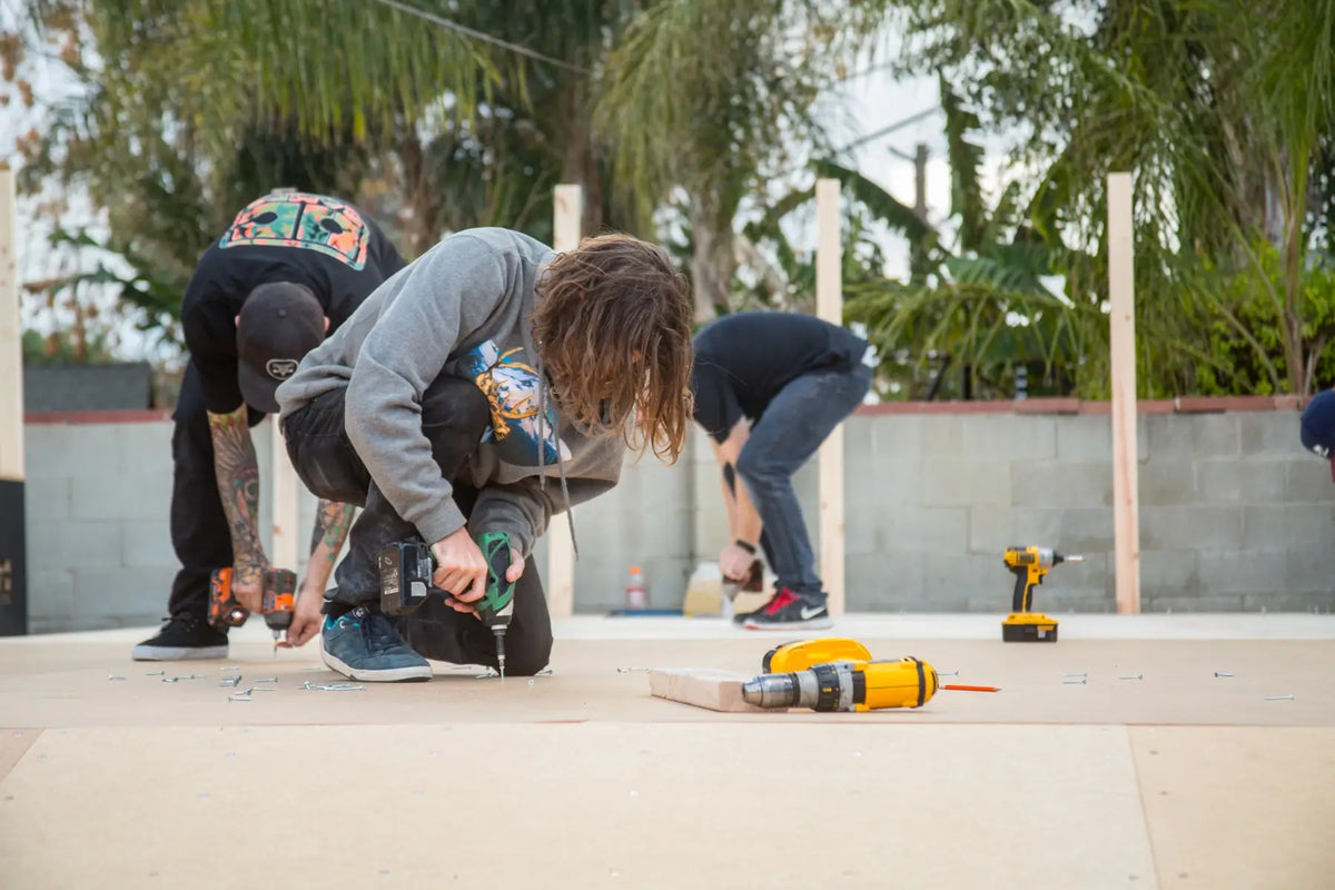 Three people working outdoors on a concrete surface, using cordless drills. A yellow and black cordless drill lies on the concrete in the foreground, with palm trees and a cinderblock wall in the background.