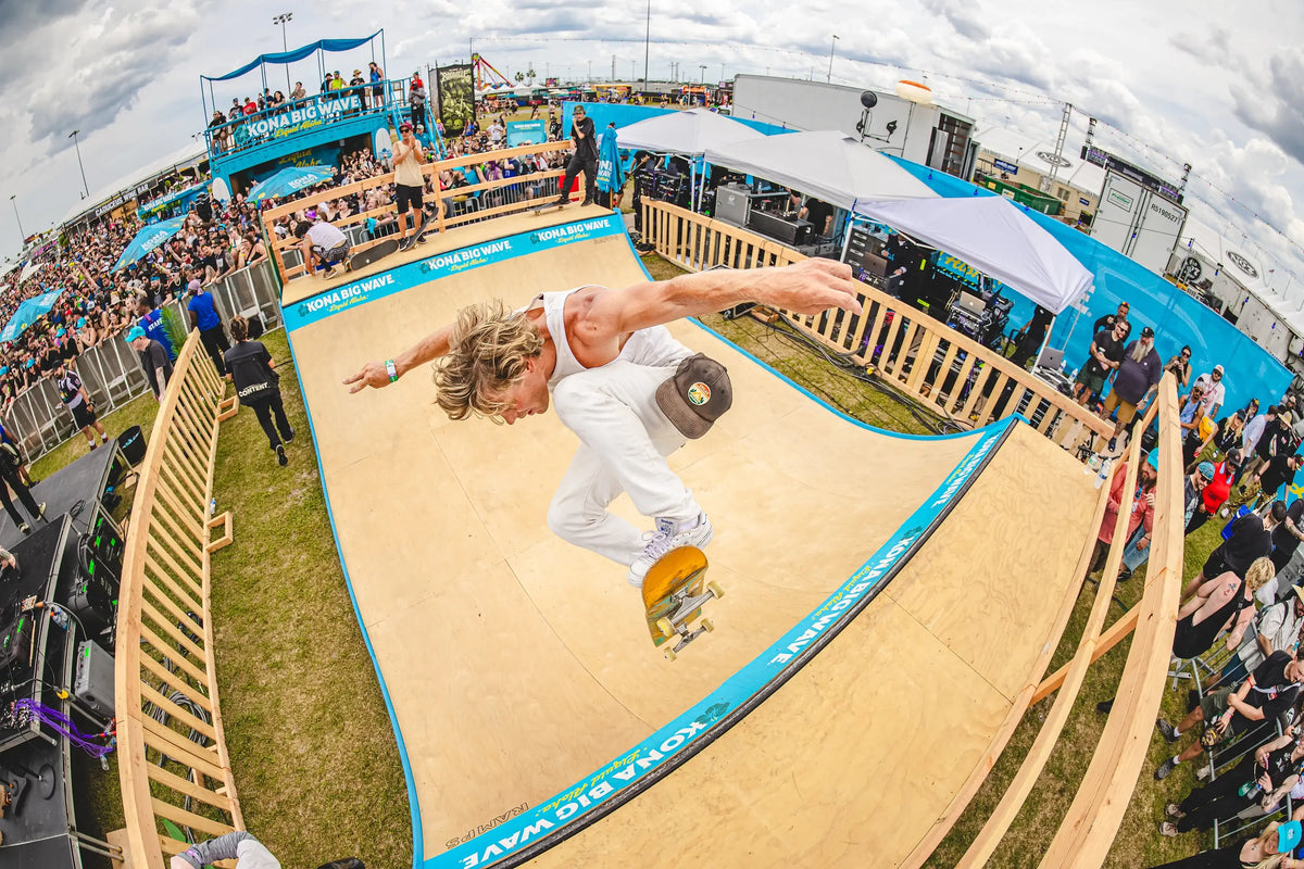 A skateboarder in white clothing performs a trick on a wooden halfpipe ramp at a Santa Monica Pier event