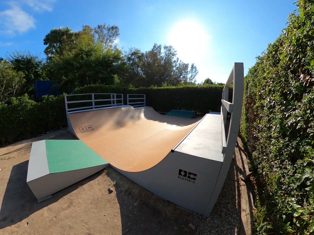 Outdoor skatepark ramp with a curved wooden bowl or quarter-pipe structure, concrete walls, a green-and-white ramp section, and surrounding hedges under a bright sun.