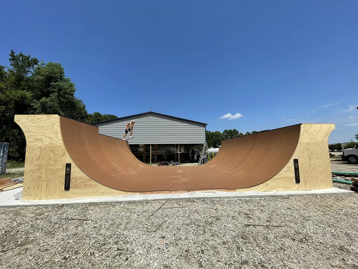 A large wooden half-pipe skate ramp constructed from plywood, photographed outdoors in a gravel lot with a metal workshop building and trees in the background under a clear blue sky.