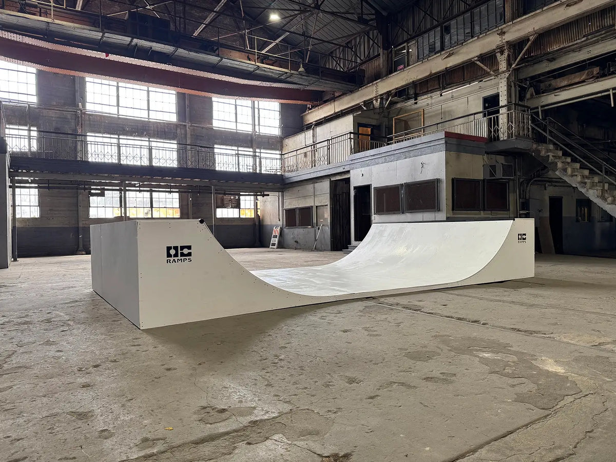 A white OC Ramps halfpipe skate ramp inside a large, industrial warehouse with concrete floors, high windows, and metal staircases.