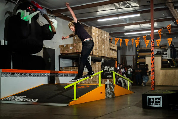 A skateboarder in a black shirt and jeans performs a trick on an orange and black ramp with a bright yellow rail inside OC Ramps' event space