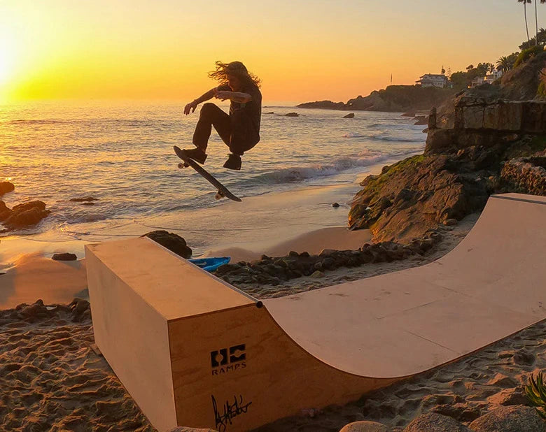 A skateboarder jumps over a wooden OC Ramps half-pipe ramp on a sandy beach at sunset, with ocean waves and rocky coastline in the background.